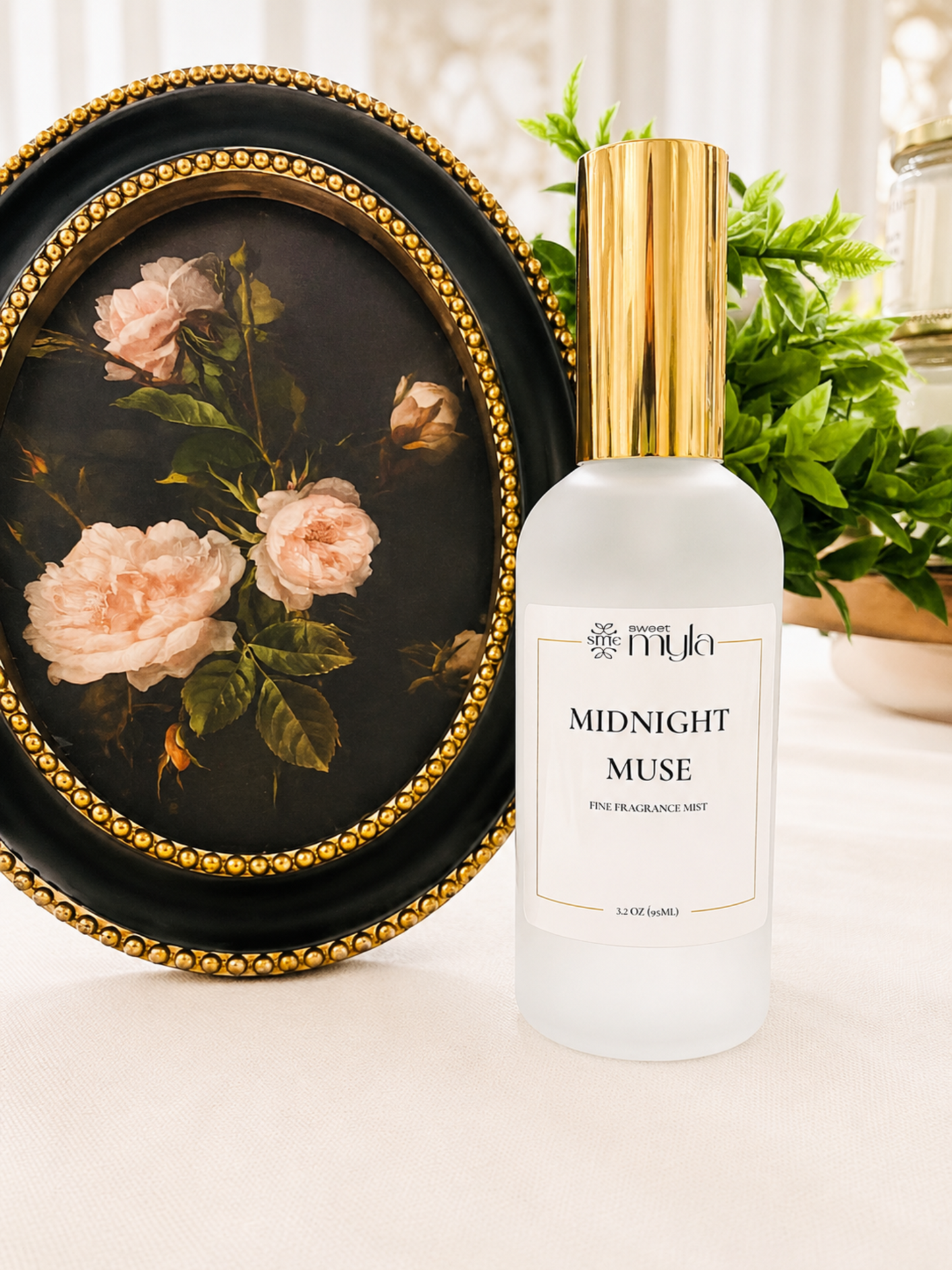 A frosted glass bottle of Sweet Myla Midnight Muse fine fragrance mist spray with a gold cap, styled next to an oval framed floral painting on a light beige surface.
