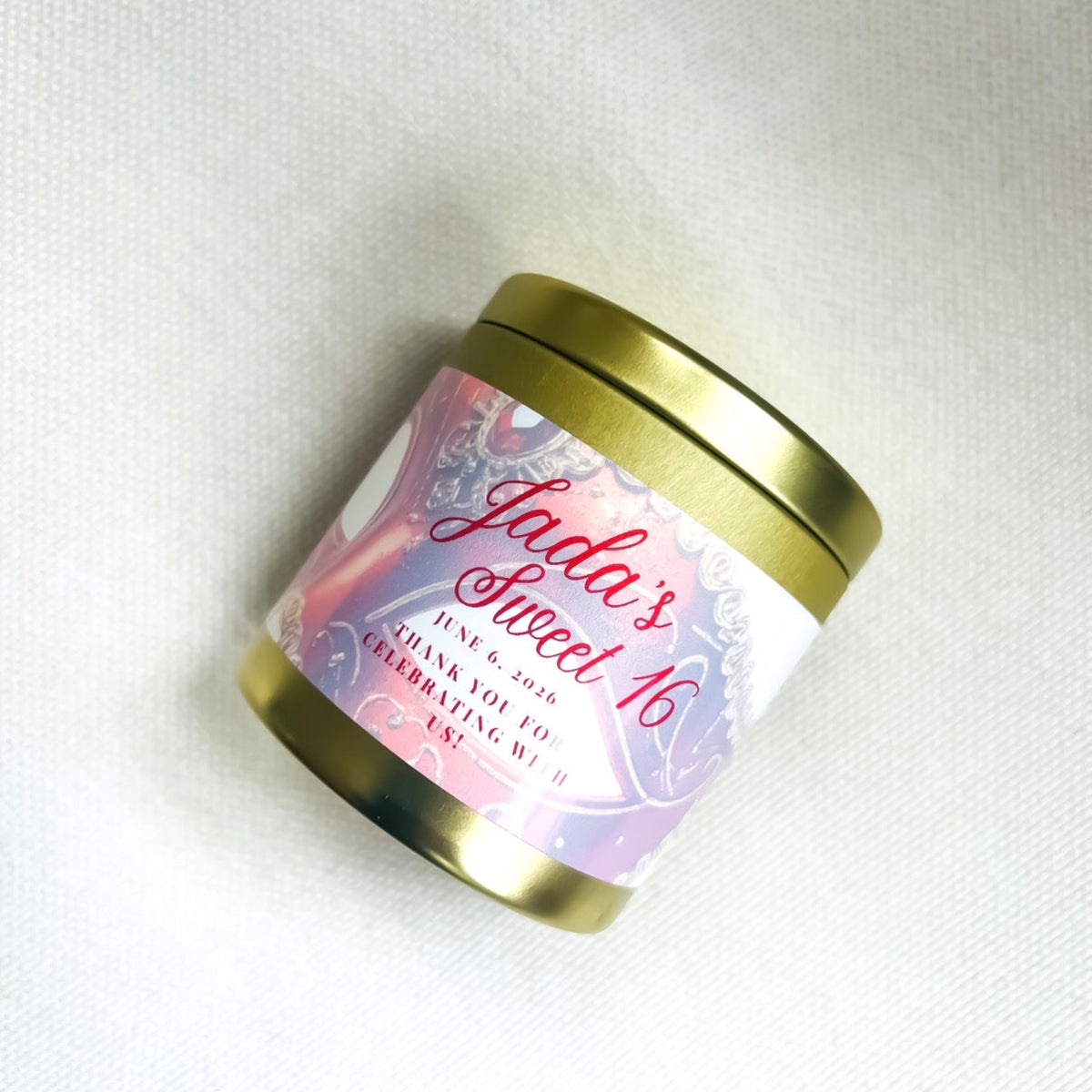 A small 4oz tin candle with a gold metal lid and base, featuring a white wrap label with red lettering and a mask artwork and the text 'Jada's Sweet 16' and 'THANK YOU FOR CELEBRATING WITH ME.' The tin is photographed on a light fabric.background.