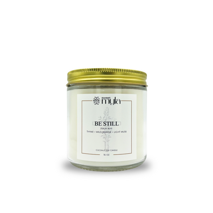 Candle jar with a gold lid labeled 'Be Still' on a white background