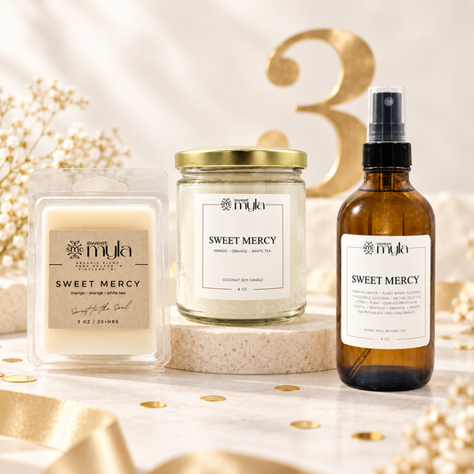  Sweet Mercy scented products arranged on a light marble surface with decorative gold ribbon, gold confetti, and a large gold number 3 in the background: a wax melt in a clear clamshell, a 8oz glass jar candle with a gold lid, and an amber spray bottle with a black pump. All products have white labels with black text in celebration of Sweet Myla 3 year anniversary. 