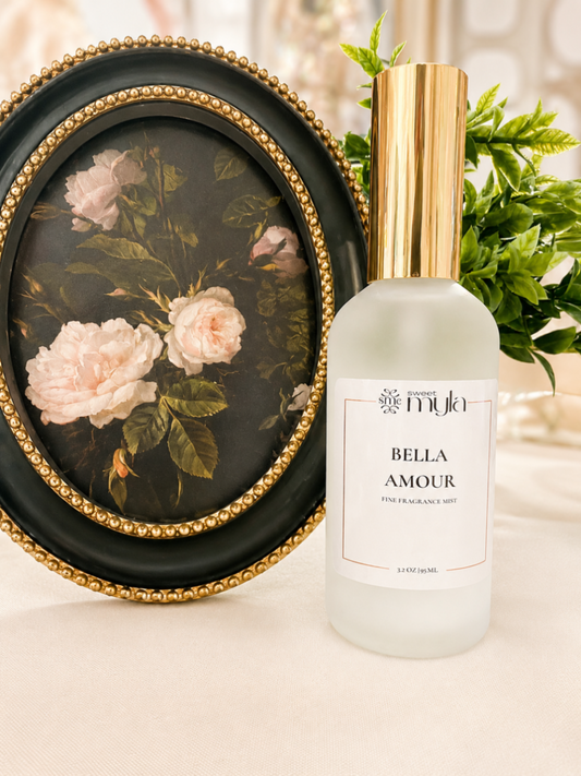 A frosted glass perfume bottle with a metallic gold cap and a white label reading 'Sweet Myla  BELLA AMOUR', styled next to an oval framed floral painting on a beige surface with green leaves in the background.