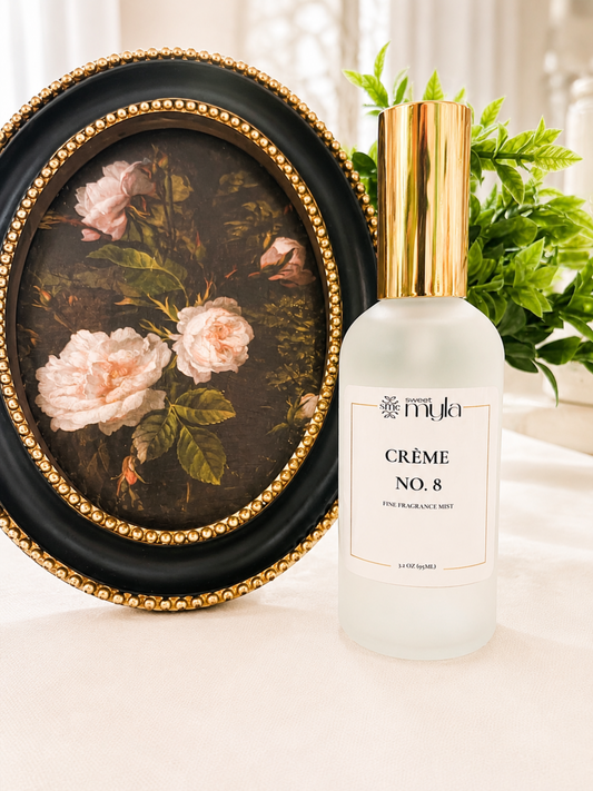 A frosted glass bottle of Myla Crème No. 8 Fine Fragrance Mist with a shiny gold cap, styled next to an oval framed floral painting on a white surface.