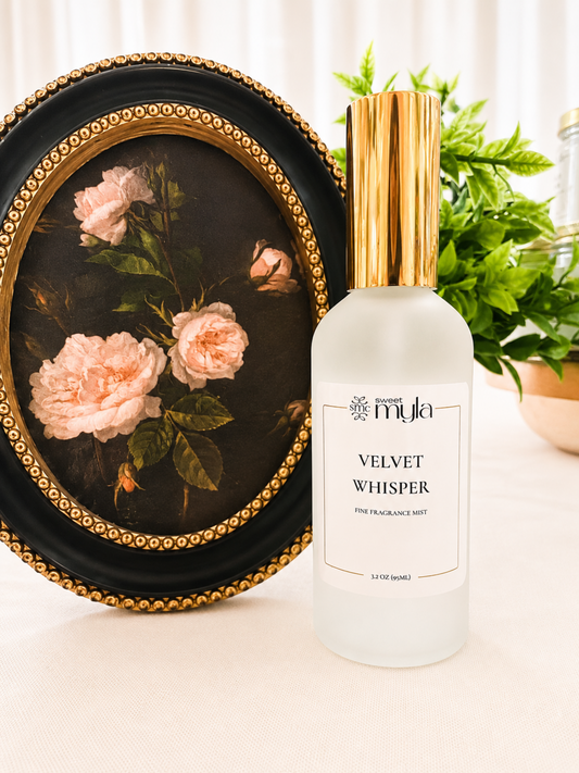 A frosted glass perfume bottle labeled 'Sweet Myla VELVET WHISPER' with a metallic gold cap, set next to a framed floral painting on a light beige surface with a green plant in the background.