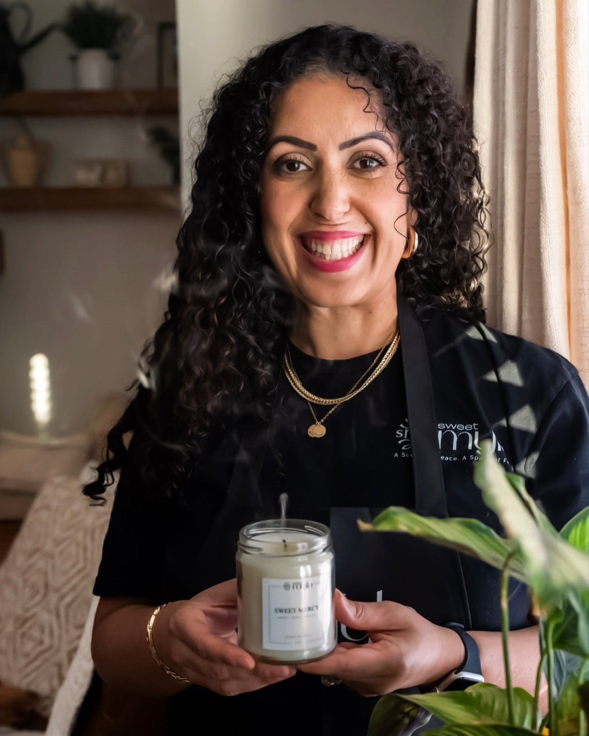 Yadi, founder of sweet Myla  with curly dark hair holds a glass jar candle in a cozy indoor setting. She is wearing a black t-shirt and apron with a white logo that reads sweet mla 