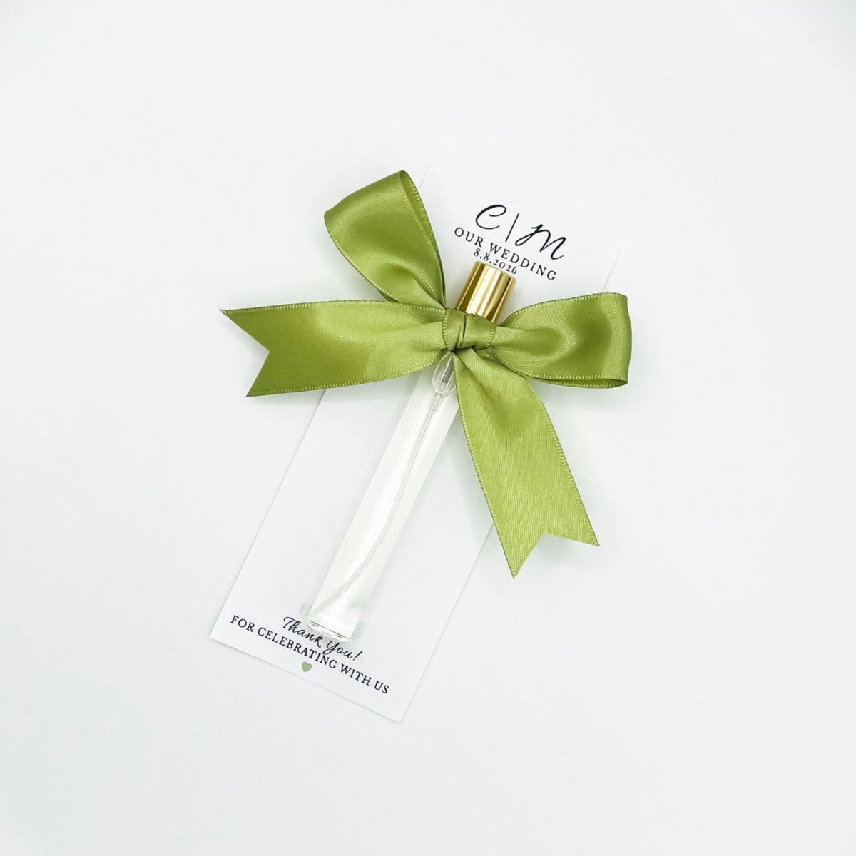 A 10ml clear glass perfume bottle tied with a wide olive green satin ribbon and a gold cap, attached to a white gift tag printed with 'C&M Our Wedding' and date. 'Thank you for celebrating with us', on a clean white background. For a wedding favor. 