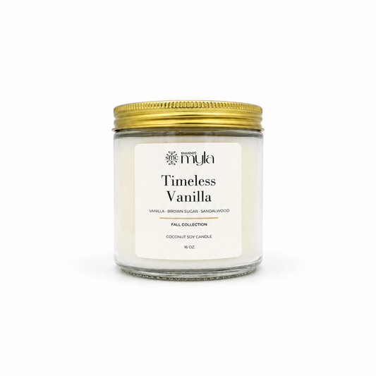 A 8 oz coconut soy candle in a clear glass jar with a white label reading 'Timeless Vanilla' by the brand 'Sweet Myla', topped with a metallic gold screw-on lid, set against a plain white background.